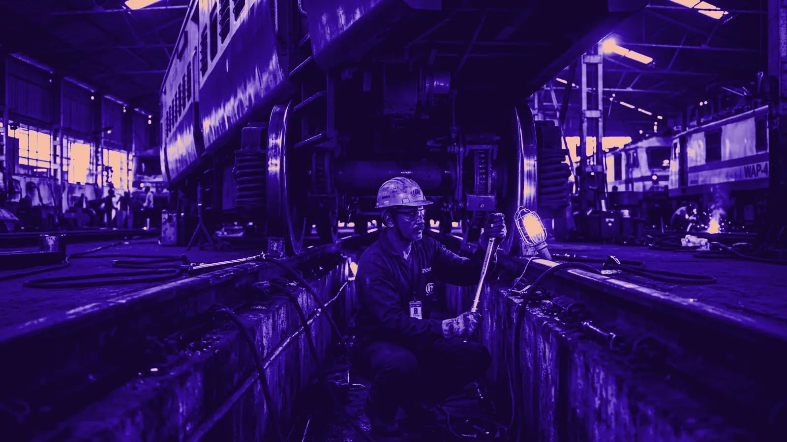 Railway mechanical technician with wrench inspecting carriage wheel bogie in a maintenance shed pit under coach chassis under workshop floodlights